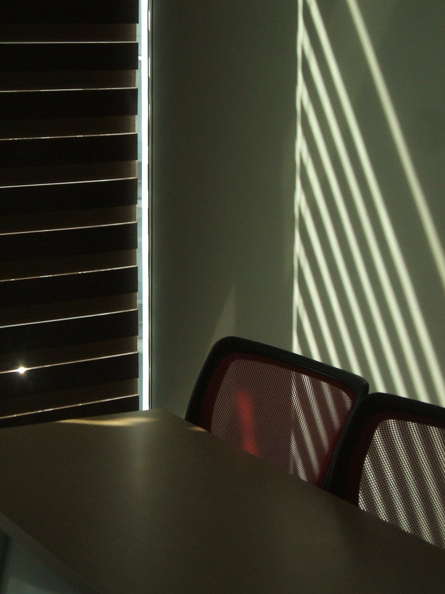 Sunlight casting dramatic shadows through blinds onto office chairs and table.