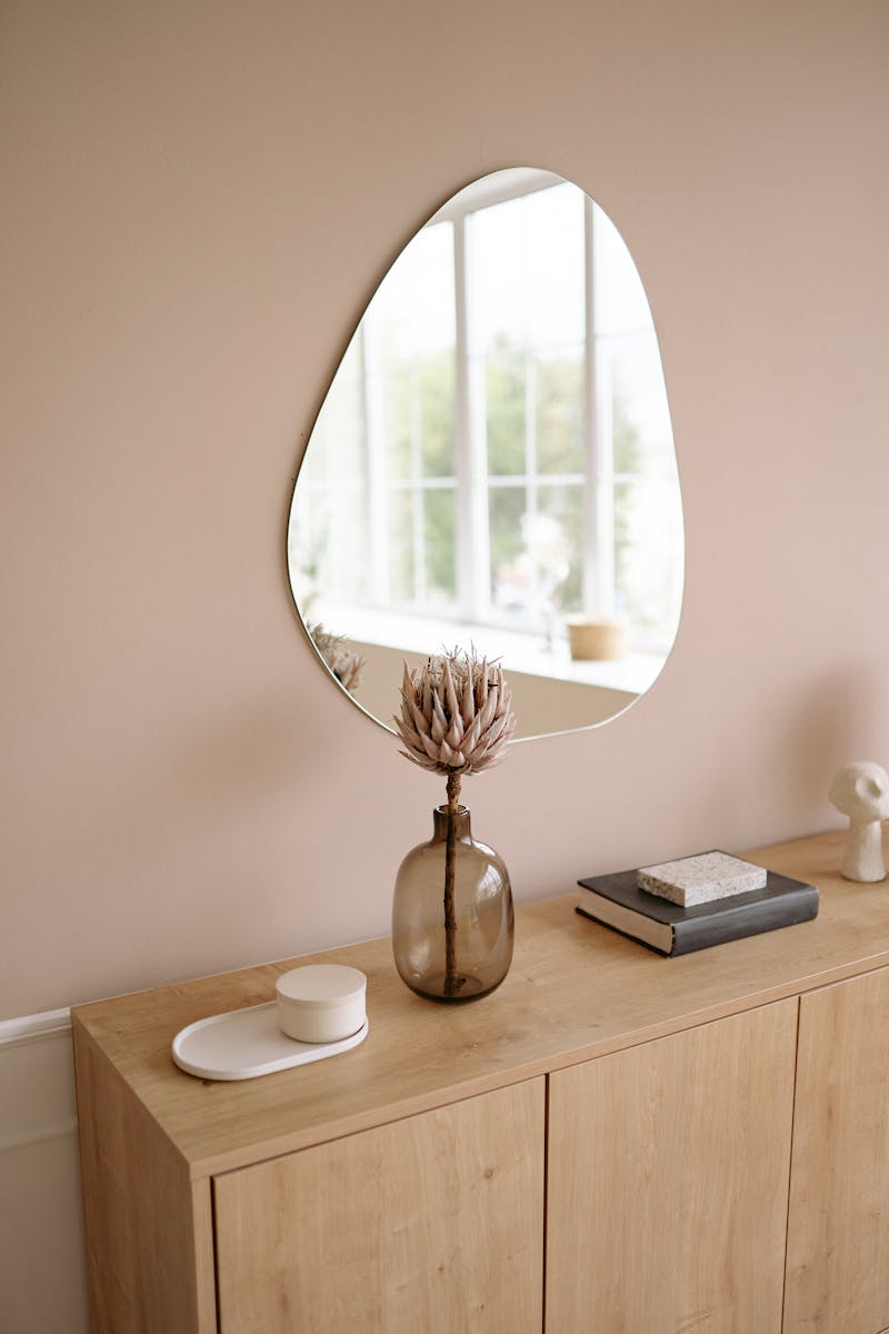 A stylish modern interior featuring a unique mirror, vase, and wooden cabinet with natural light.